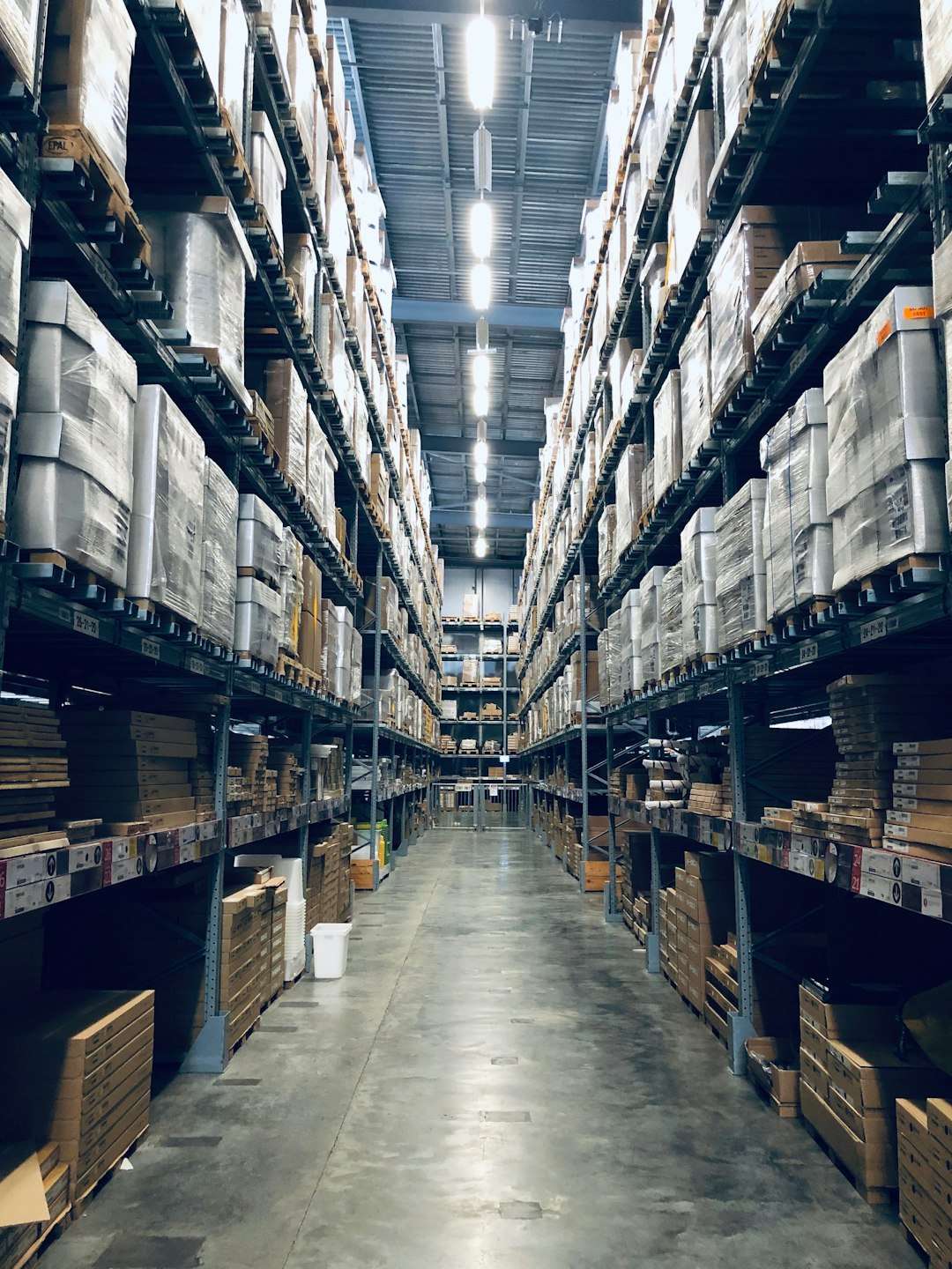 Long warehouse aisle with tall metal shelves packed with stacked pallets and boxes, illuminated by overhead lights.