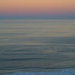 Silhouette of a person walking along a dark rocky shore with a calm sea and pastel sunset sky on the horizon.