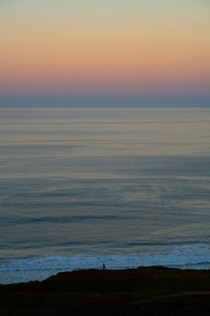 Silhouette of a person walking along a dark rocky shore with a calm sea and pastel sunset sky on the horizon.