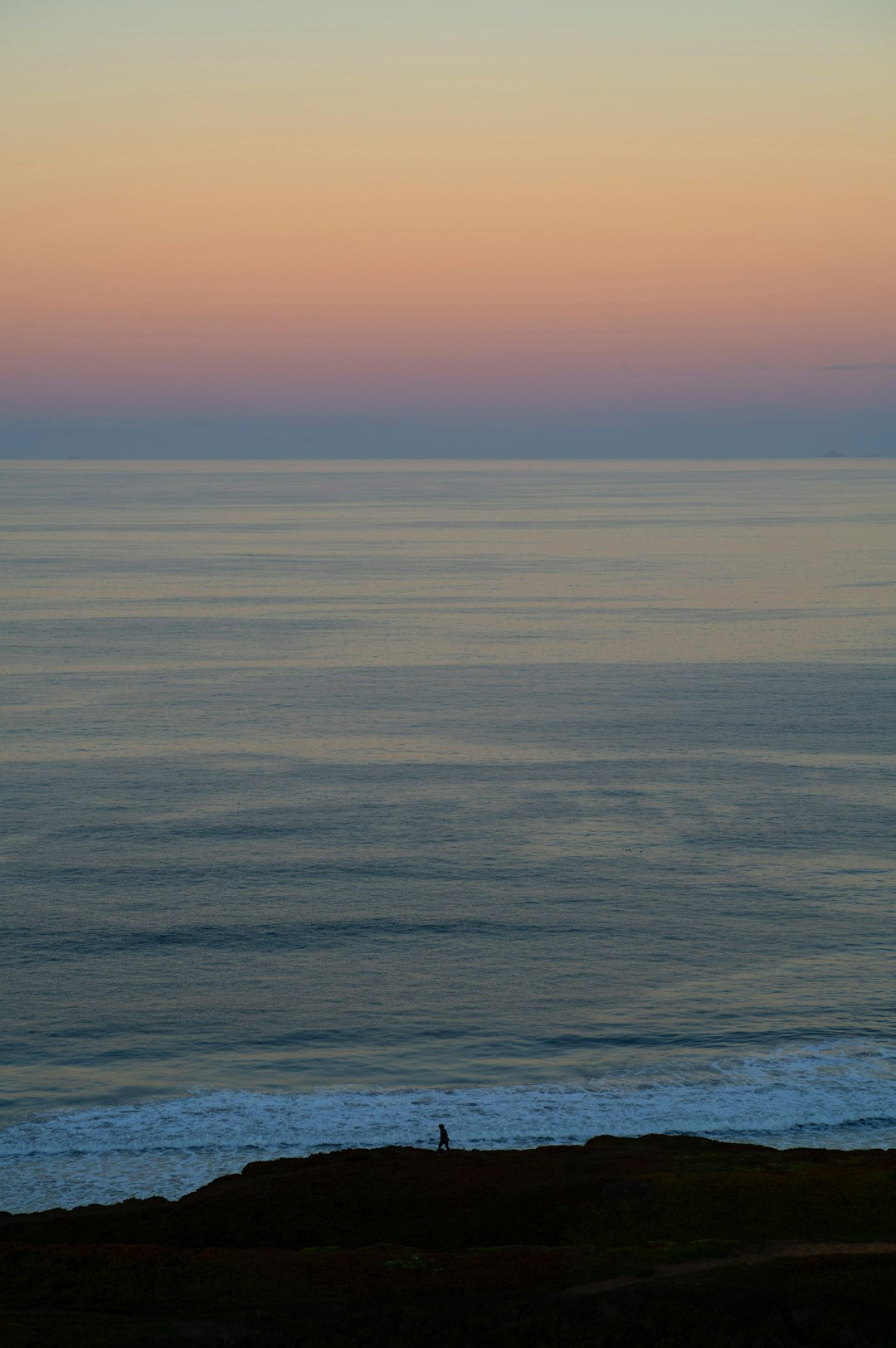 Silhouette of a person walking along a dark rocky shore with a calm sea and pastel sunset sky on the horizon.