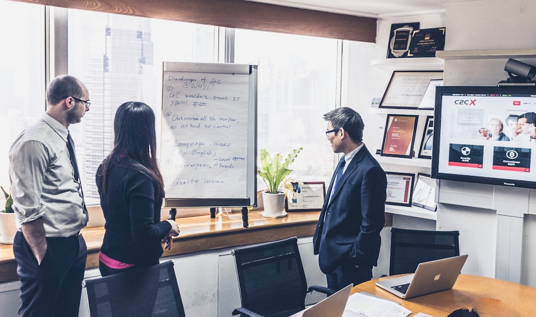 Three professionals discuss ideas around a whiteboard with handwritten notes in a bright office overlooking a city skyline.
