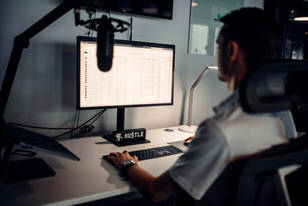Person sits at a desk with a computer monitor and a large overhead microphone, likely recording or streaming.