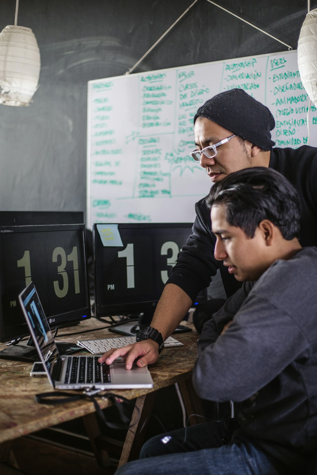 Two people work together at a wooden desk with laptops and large clock-style screens, whiteboard full of notes behind them.