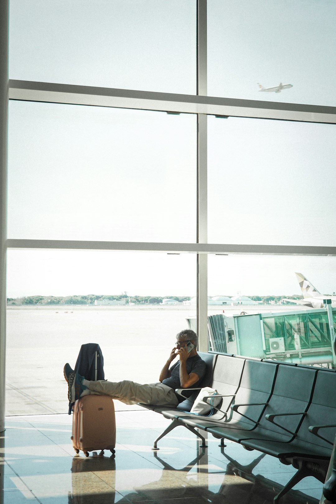 Man reclining in an airport chair with a suitcase beside him, talking on his phone by large windows.