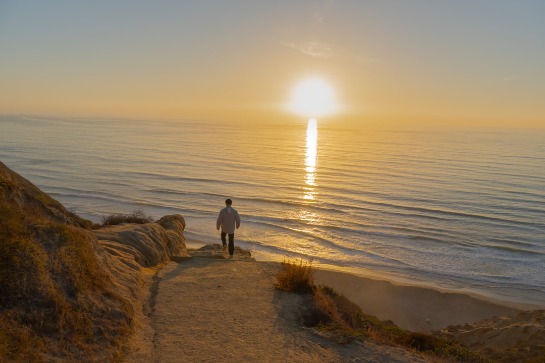 Person walking down a sandy path toward a sunlit beach as the sunset reflects on the calm ocean waves in front of them with a golden sky.