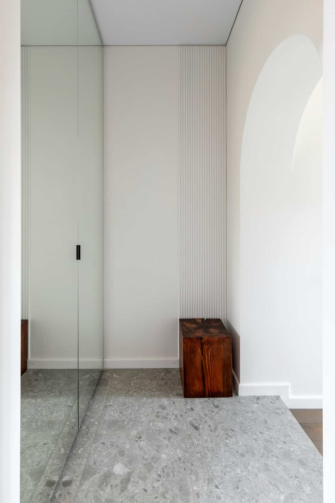 White minimalist hallway corner with a tall built-in cabinet that has a mirrored door and a ribbed wall detail; a wooden block stool sits on a grey terrazzo floor.