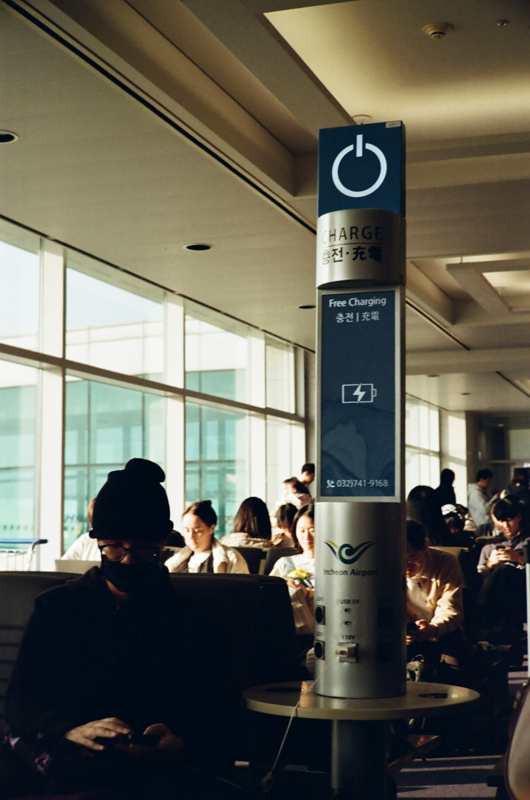 Charging station pillar at an airport terminal with a 'Free Charging' sign; passengers seated in background.