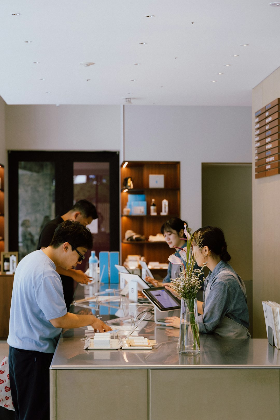 Reception desk with staff assisting customers at a modern counter, tablet screens in use, and a vase of flowers in the foreground.