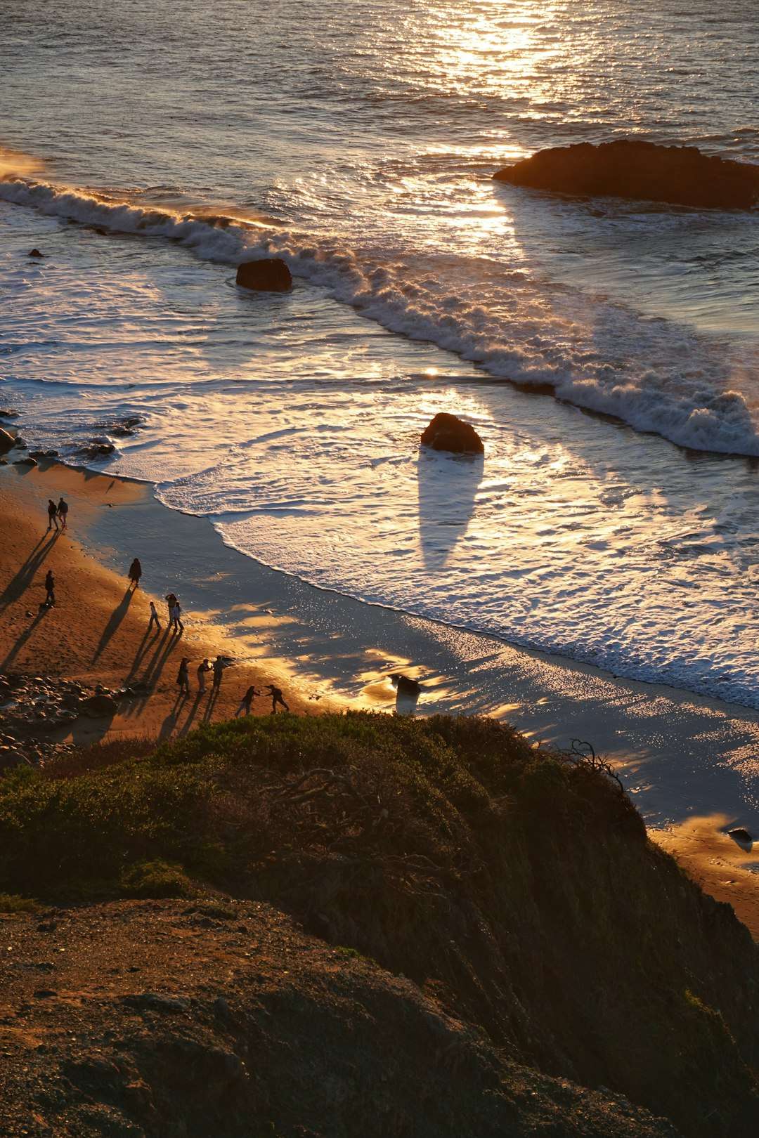 Sunset over a rocky beach with golden light reflecting on waves and several silhouetted people walking along the shore.