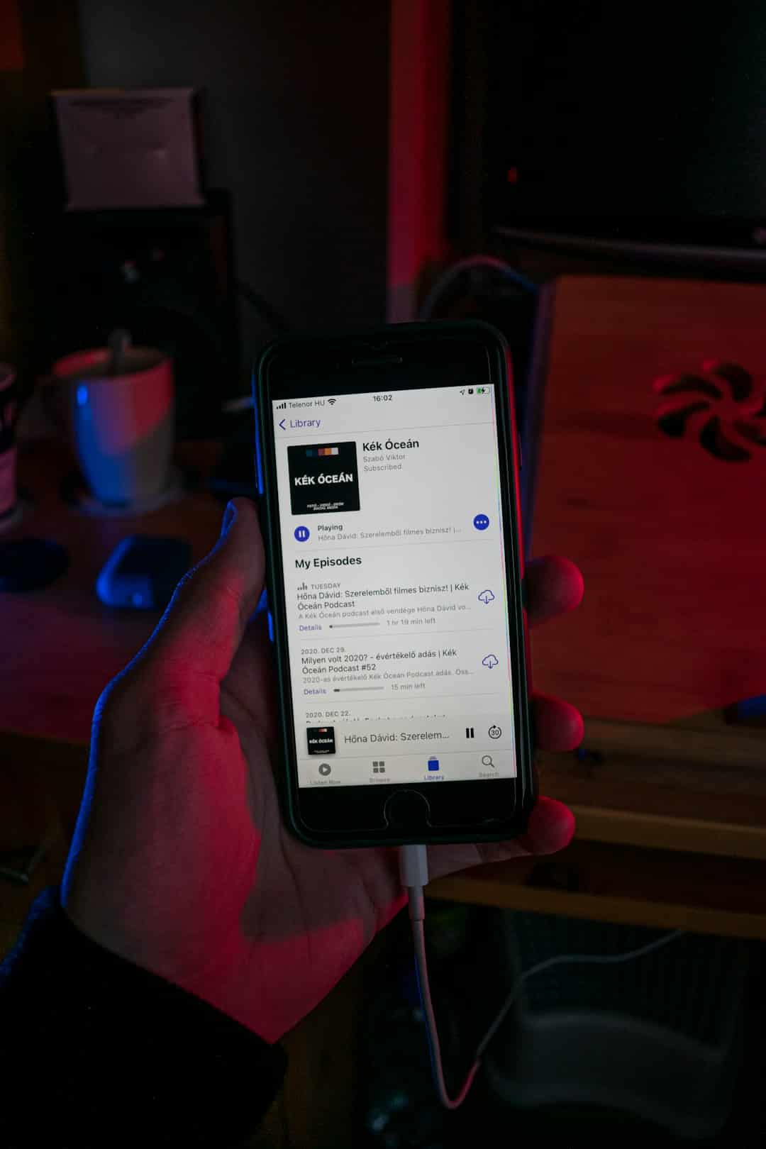 Hand holding a phone in a dimly lit room, showing a podcast app with 'Kék Óceán' in the library view