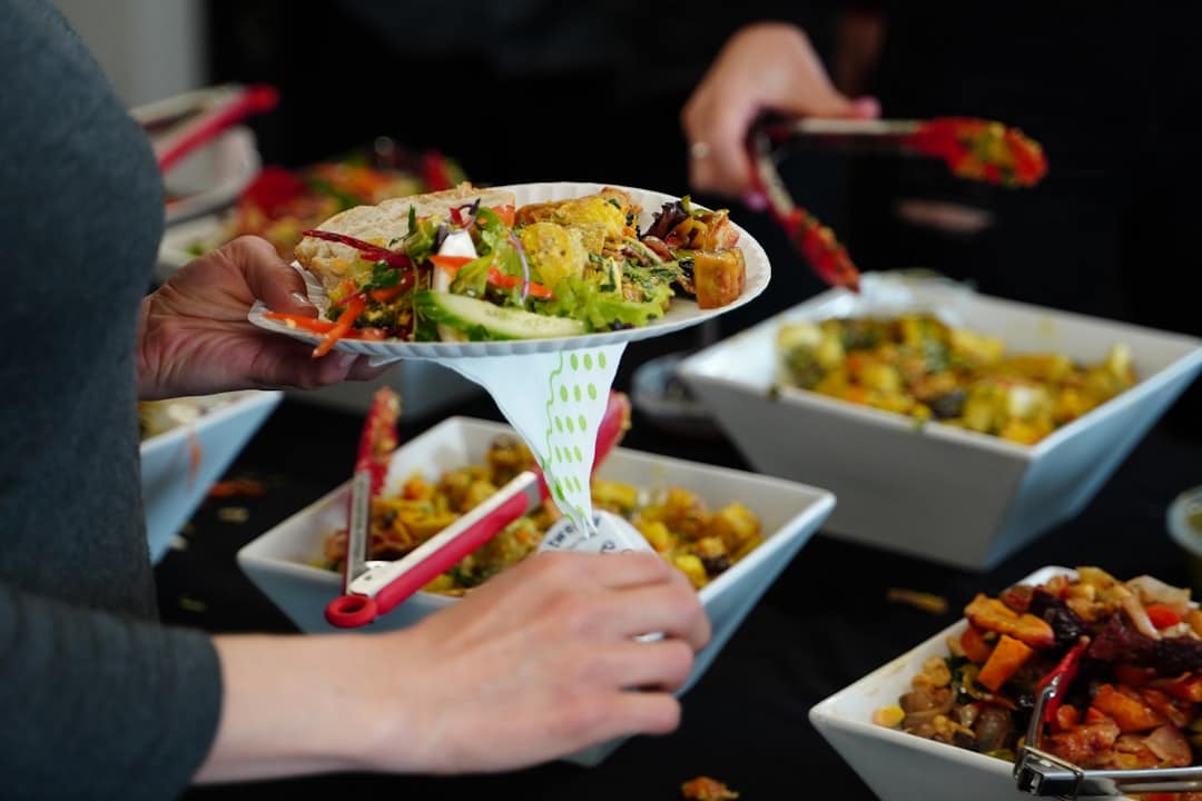 Person serving a plate of colorful salad and vegetables from a buffet, with other dishes in white bowls on a dark tablecloth behind them