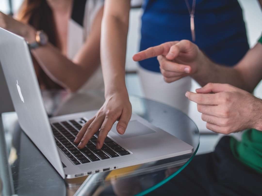 Hands typing on a silver laptop at a glass table while two colleagues point and discuss nearby.