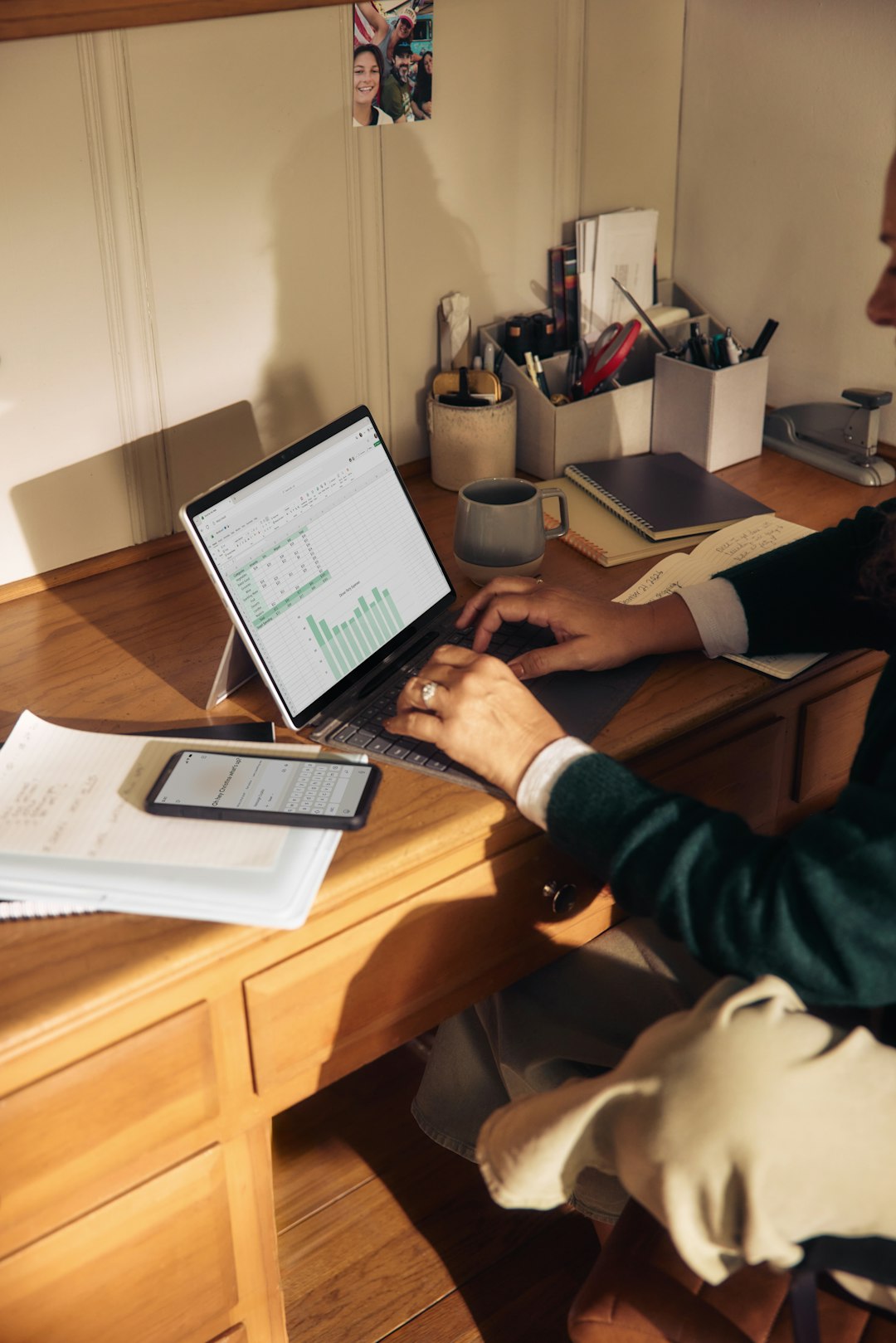 Person typing on a tablet-laptop at a wooden desk, with charts visible on the screen and a phone nearby.