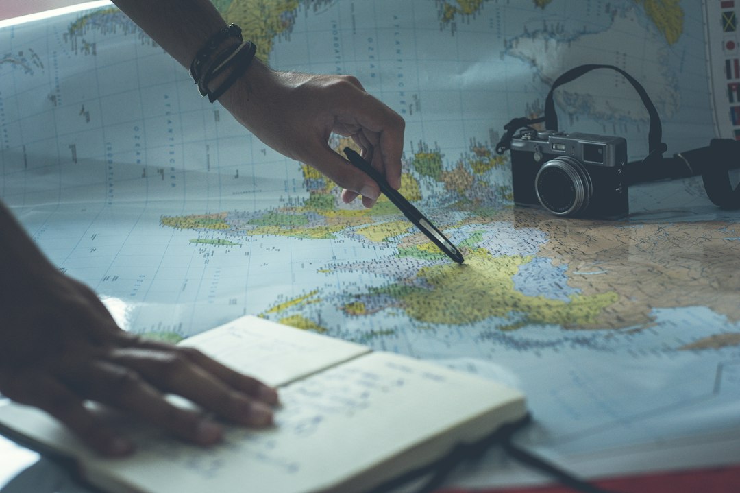 Person using a stylus to point at a world map, with a notebook and a camera resting nearby for travel planning.