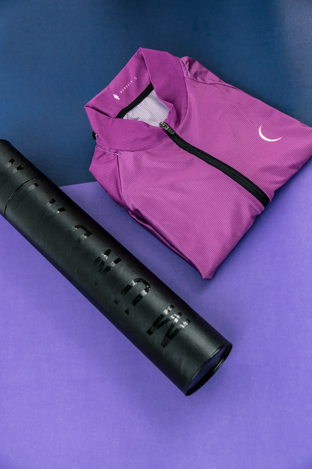 Folded purple zip-up jacket with a black zipper beside a black water bottle on a two-tone purple background.