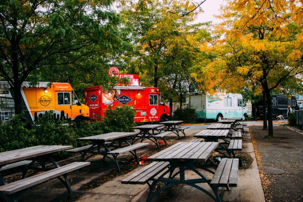 Row of wooden picnic tables in a wet parking lot with colorful food trucks and fall foliage in the background.