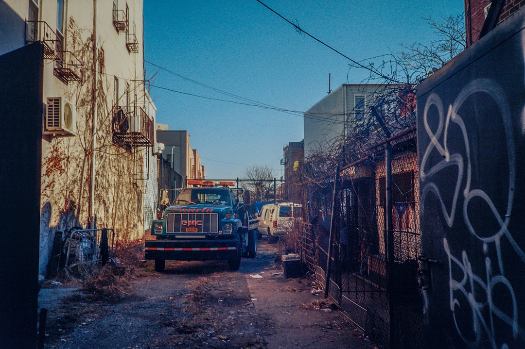 Blue GMC tow truck parked in a narrow alley between graffiti-covered fences and brick buildings behind it.