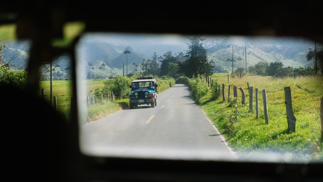 Blue vintage jeep driving toward the camera on a narrow rural road, flanked by green fields and wooden fences with mountains in the distance.