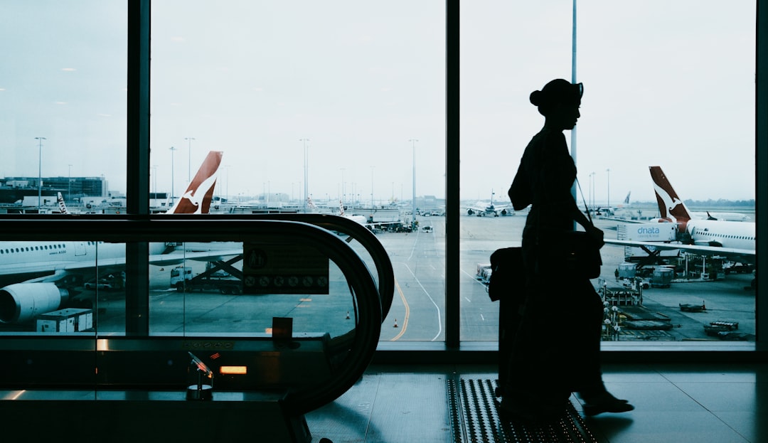Silhouette of a traveler with a rolling suitcase walking past large windows overlooking a busy airport tarmac, by an escalator on the left.