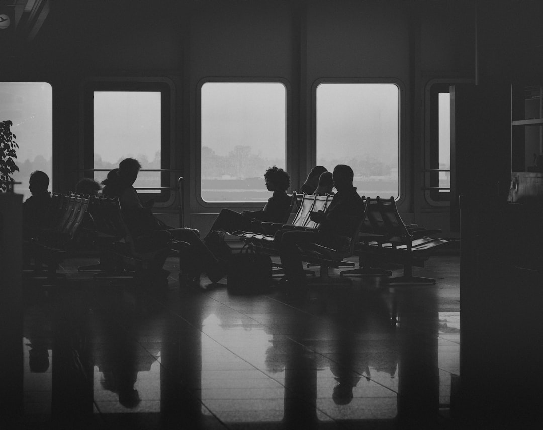 Silhouetted travelers sit in a dim transport terminal by large windows, with reflections on the polished floor.