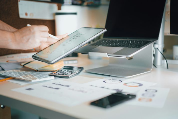 Person uses a stylus to write on a tablet propped above a laptop on a standing desk setup, with a calculator and papers nearby on the desk as reference material.