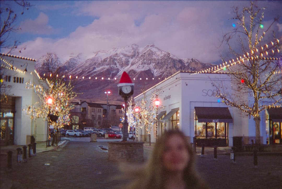 Festive shopping street at dusk with string lights, storefronts, a clock tower wearing a red Santa hat, and snow-capped mountains in the background (blurred figure in foreground).