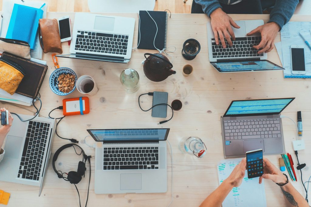 Top-down view of a busy coworking desk with multiple laptops, phones, notebooks, and coffee on a light wood table