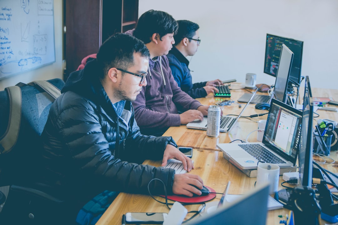 Team of developers working at a long wooden table with laptops, monitors displaying code, and a whiteboard in the background.