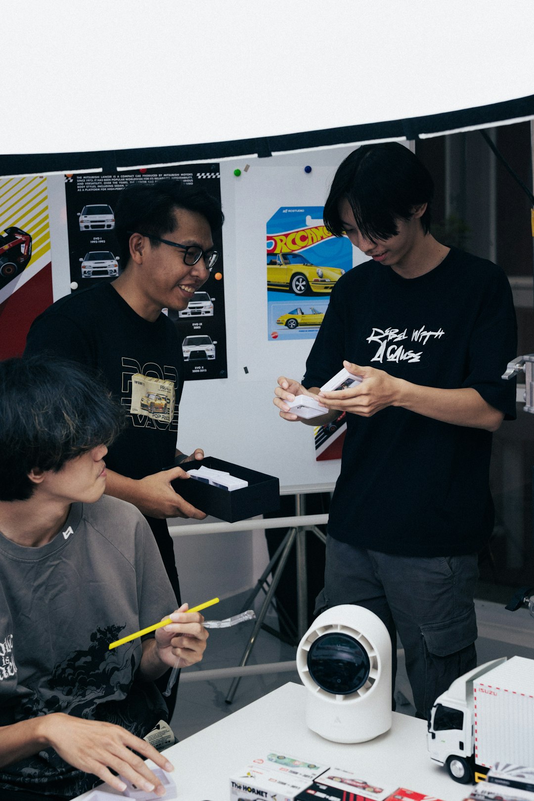 Three young men in a hobby workshop inspect and share model car kits at a table with car posters on the wall behind them.