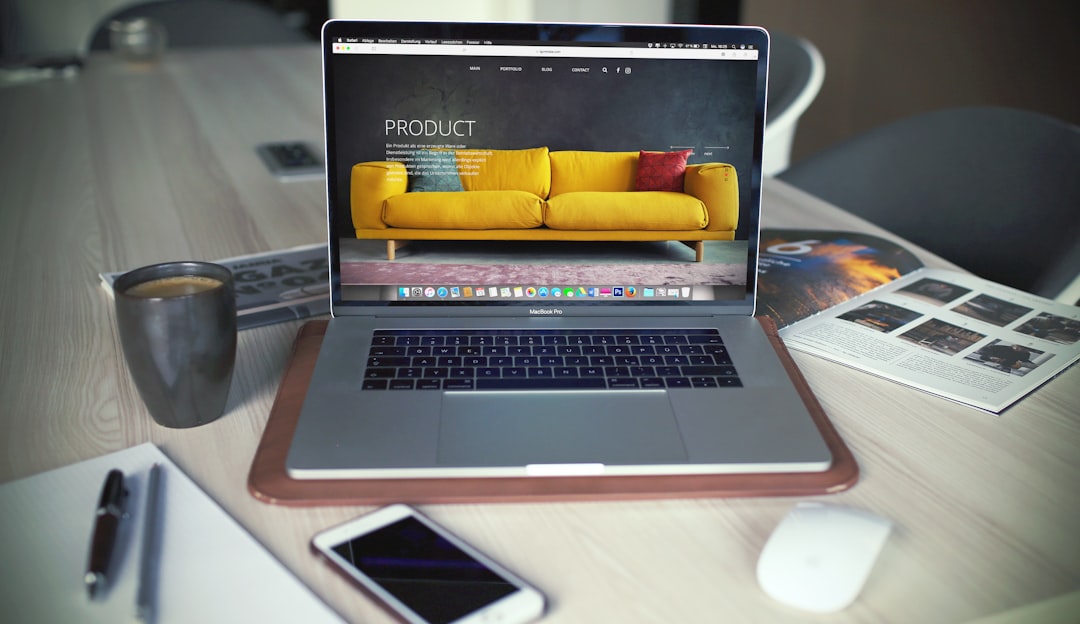 Laptop on a wooden desk showing a webpage with a bright yellow sofa image on screen, alongside a coffee mug and phone nearby, in a modern workspace.