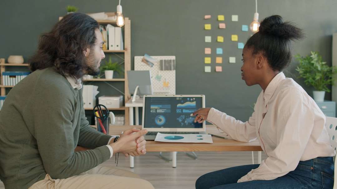 Two colleagues discuss data together, looking at a laptop with charts on a wooden table in a bright office setting.