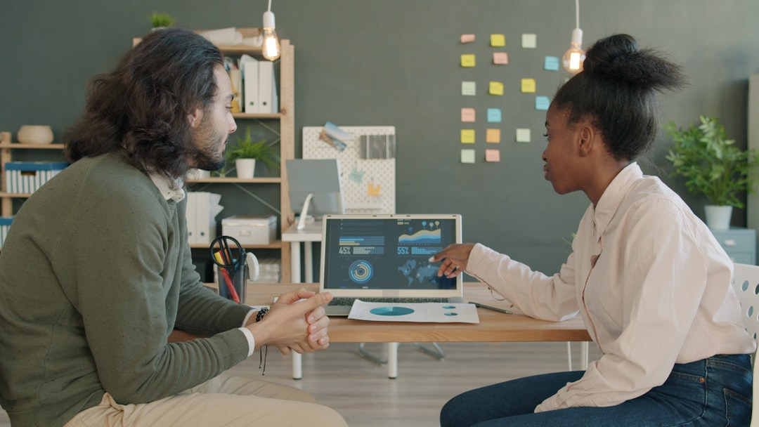 Two colleagues discuss data at a desk, one pointing to a laptop displaying charts and graphs.