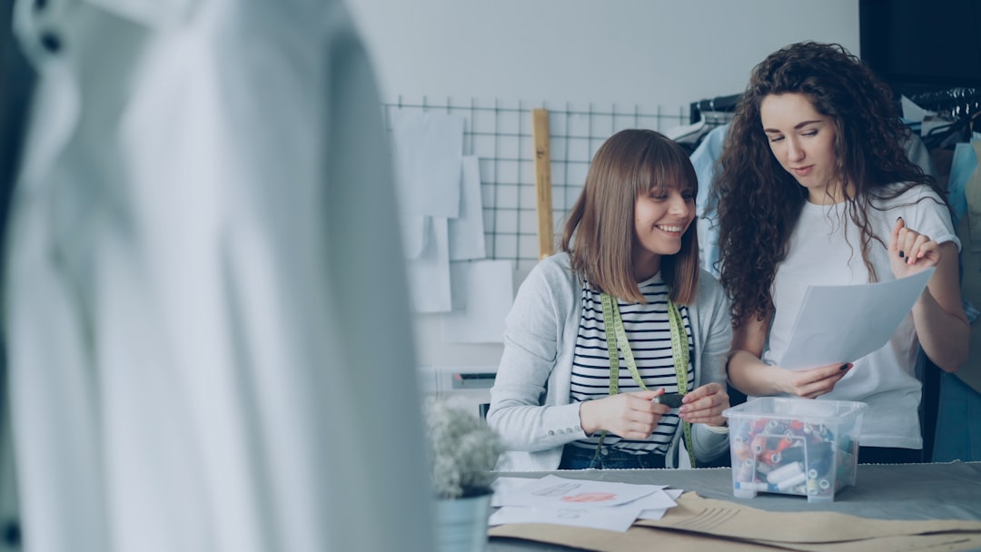 Two women in a sewing studio review patterns and papers at a table, smiling as they discuss the designs with sewing supplies nearby