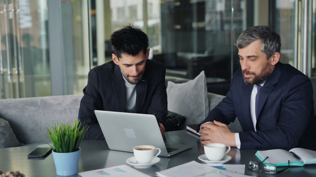 Two men in suits collaborate at a glass-topped table, focusing on a laptop with coffee cups, a plant, and a notebook nearby in a modern office setting.