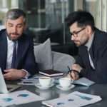 Two businessmen in a modern office review documents on a laptop and charts at a table.