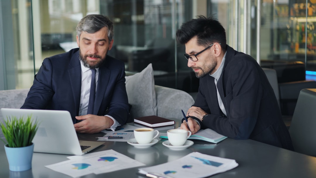 Two businessmen in a modern office review documents on a laptop and charts at a table.