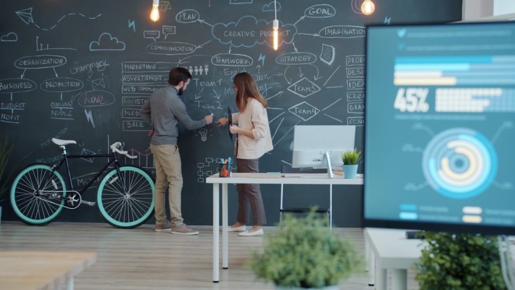 Two coworkers discuss ideas at a chalkboard filled with diagrams and notes in a creative office, with a mint bike beside them and a desk with a computer nearby.