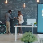 Two coworkers discuss ideas at a chalkboard filled with diagrams and notes in a creative office, with a mint bike beside them and a desk with a computer nearby.