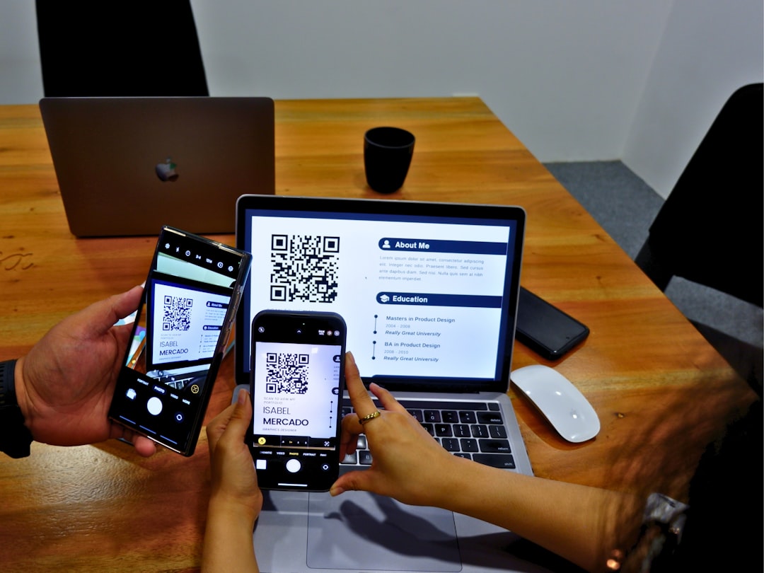People at a wooden desk use two smartphones to scan QR codes displayed on a laptop screen and a tablet device nearby.