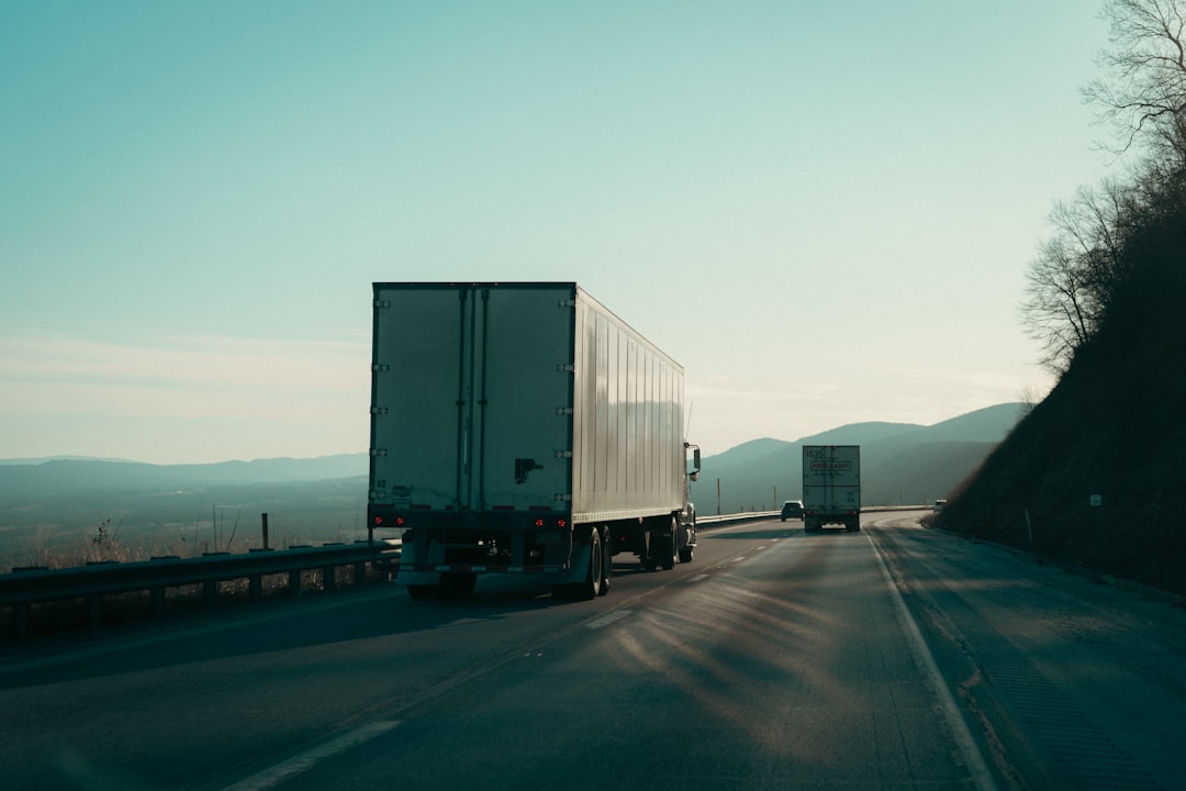 Two semi-trucks driving on a mountainous highway at dusk, with a guardrail on the left.