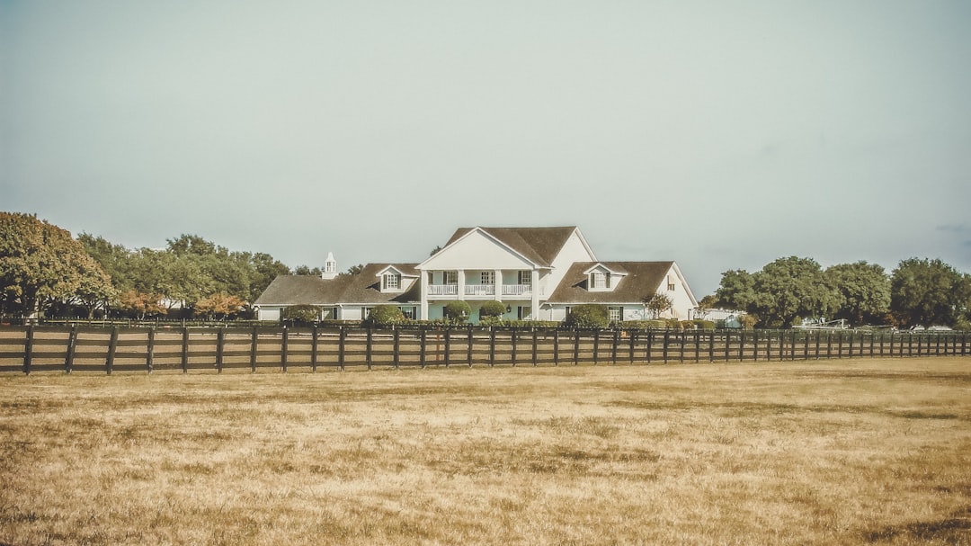 White farmhouse with a steep, multi-gabled roof behind a long wooden fence, set in a dry yellow field with trees along the horizon.
