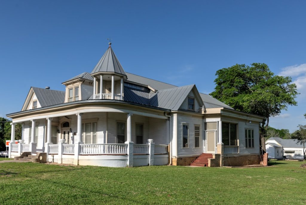 White Victorian-style house with a wraparound porch, tall columns, and a small turret under a clear blue sky on a sunny day.