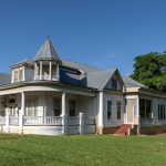 White Victorian-style house with a wraparound porch, tall columns, and a small turret under a clear blue sky on a sunny day.