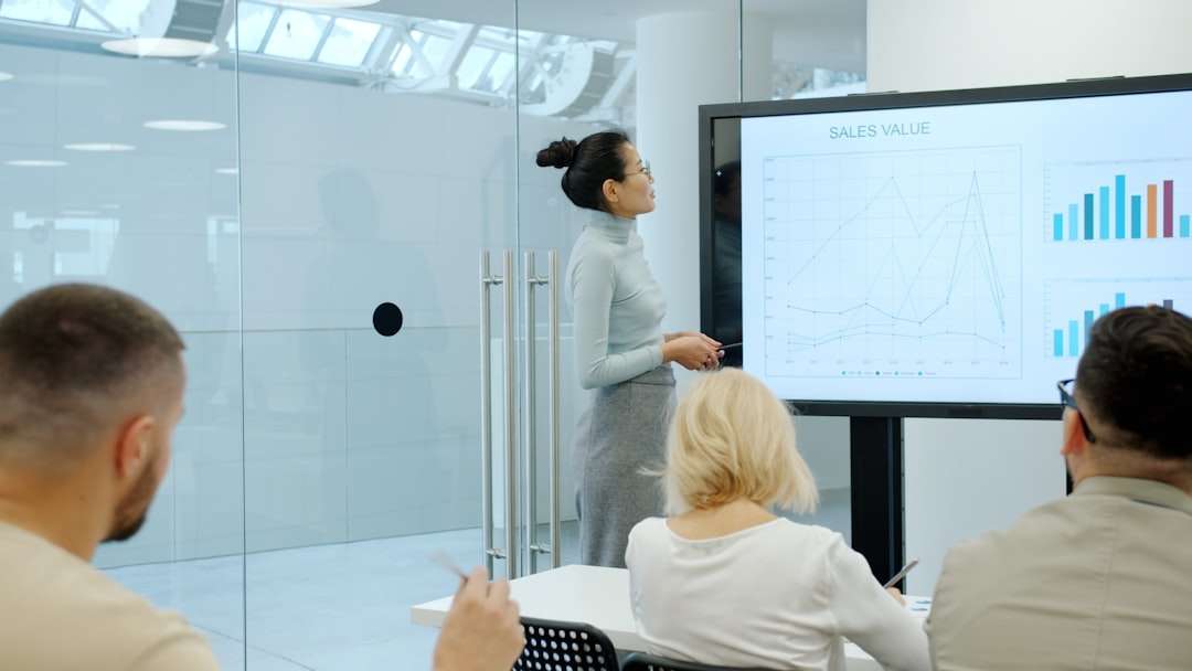 Business presentation: a woman presenter stands beside a large screen displaying sales charts to three colleagues in a glass-walled meeting room under bright lighting.