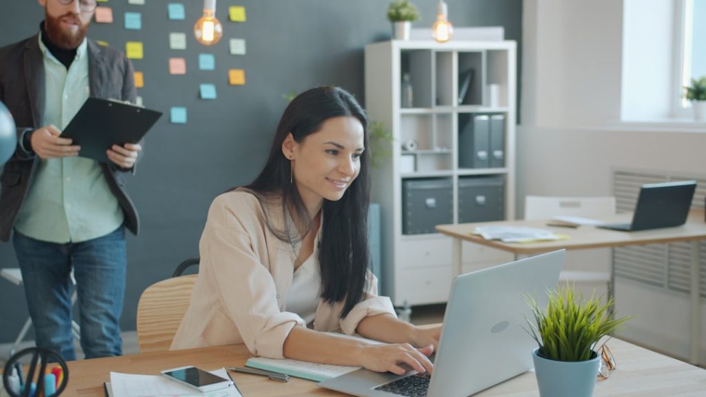 Woman typing on a laptop at a wooden desk in a bright office; a man with a clipboard stands in the background with sticky notes on a wall behind them.
