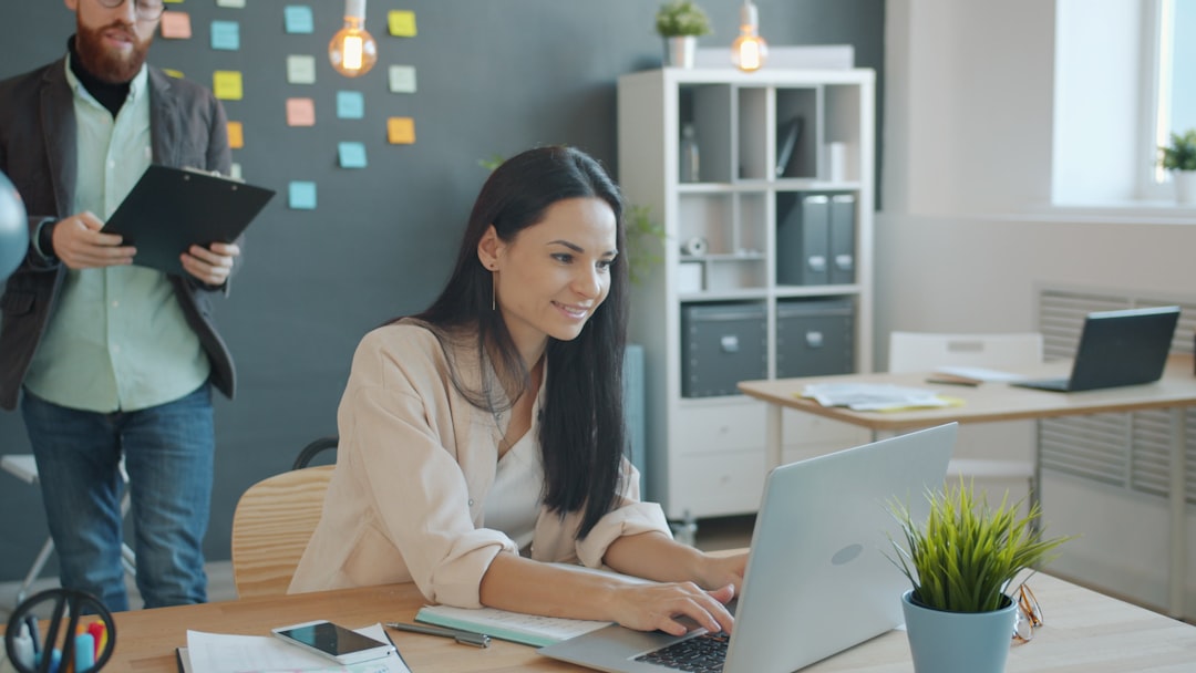 Woman typing on a laptop at a wooden desk in a bright office; a man with a clipboard stands in the background with sticky notes on a wall behind them.