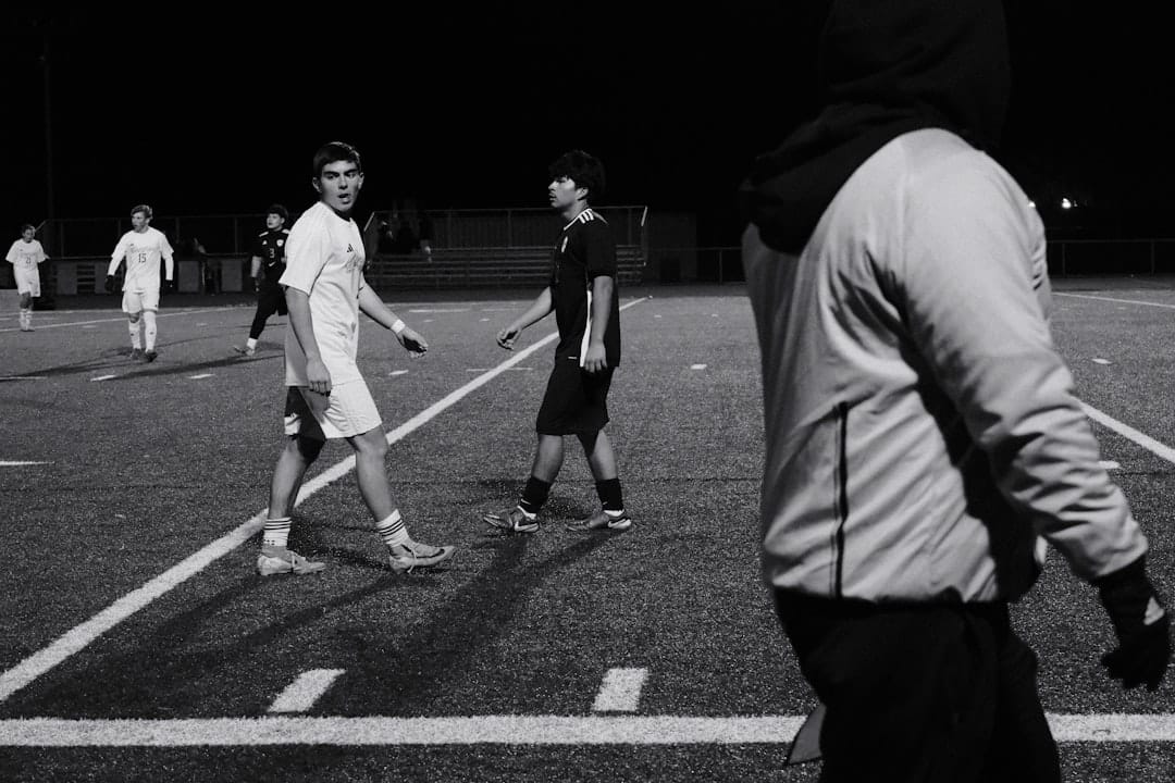 Nighttime soccer scene with several players in white and dark kits walking on a floodlit field. The foreground shows a hooded figure on the right.