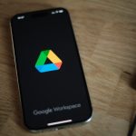 Smartphone on a wooden desk showing the Google Workspace logo on a dark screen from a slight angle.