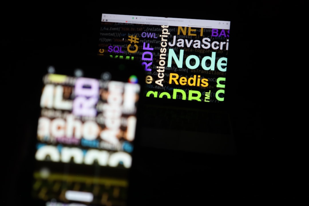 Blurred laptop screen showing colorful programming keywords (Node, Redis, JavaScript) with a foreground out-of-focus phone screen on dark background.