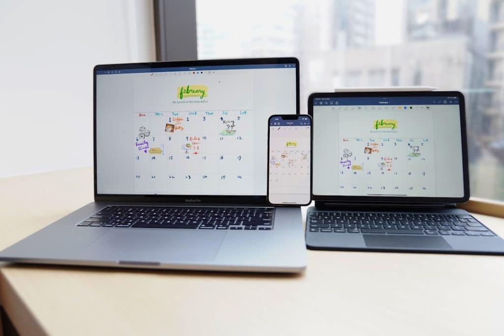 Triple-device setup showing a colorful February calendar on a laptop, tablet, and phone on a desk by a bright window